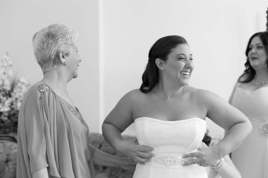 Black and white photo of bride laughing as she puts her dress on.