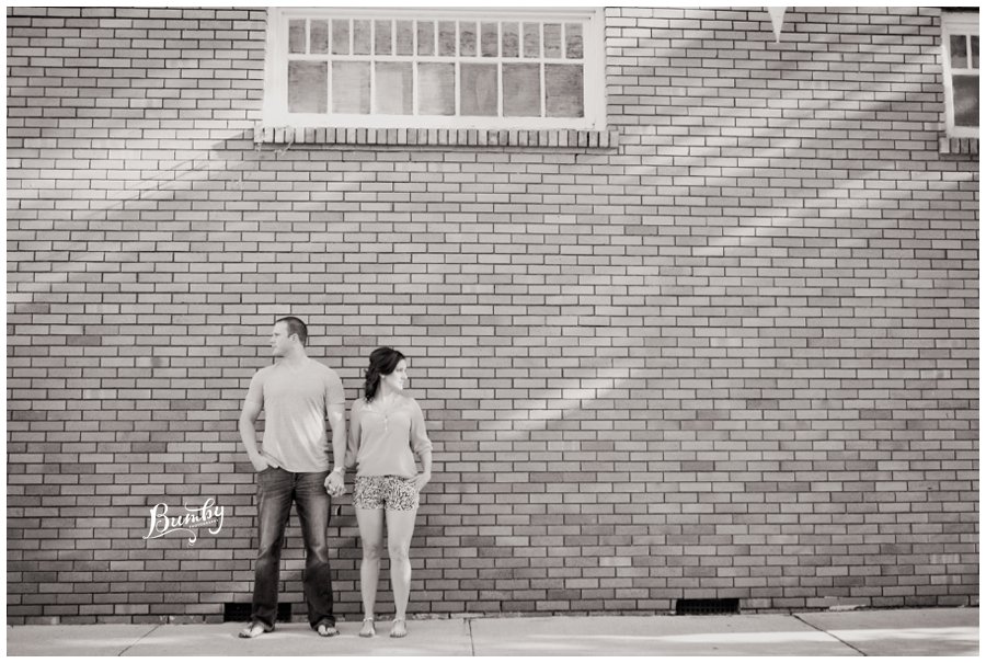 Black and white photo of a couple holding hands and facing away from each other while standing next to a brick wall.