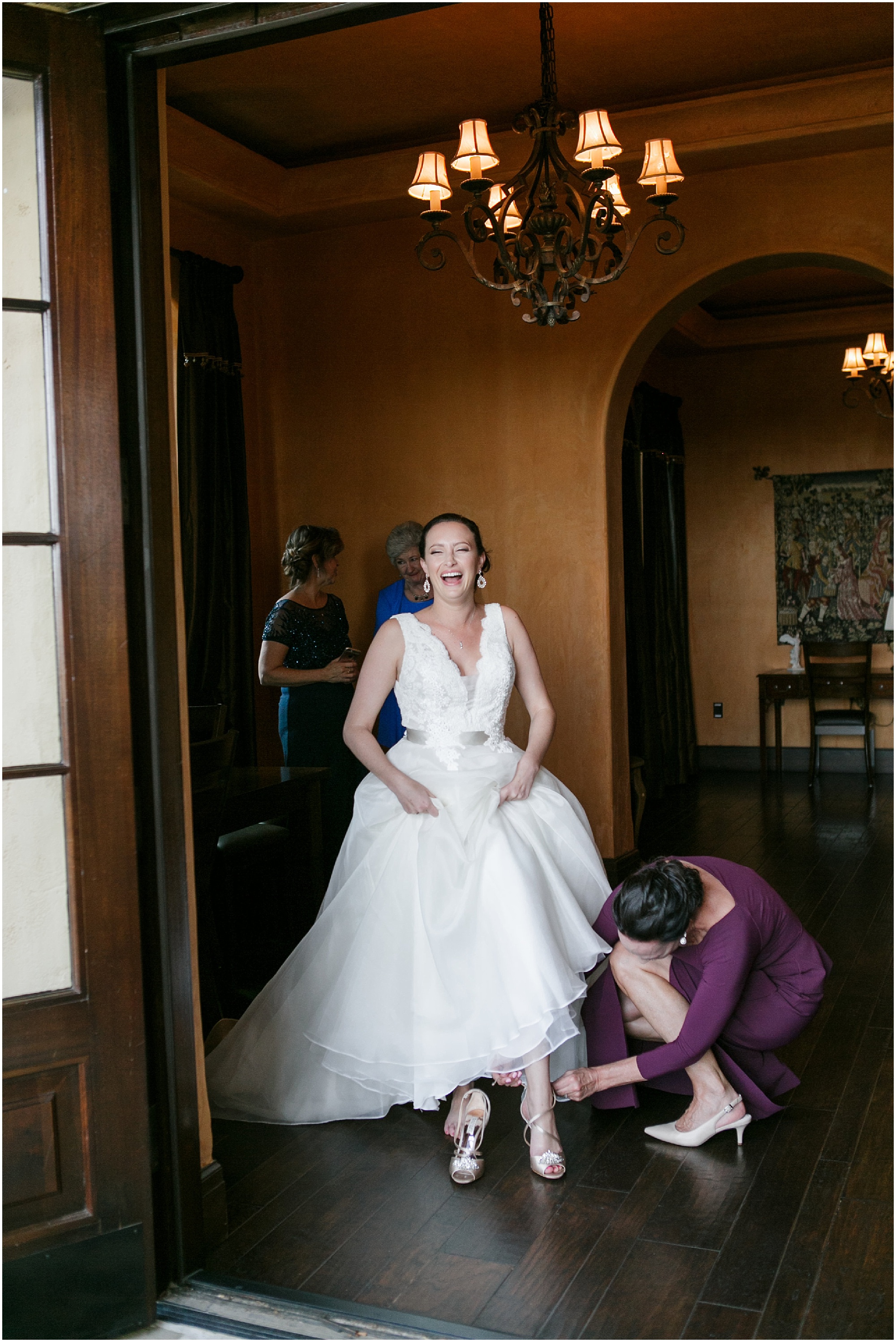 Bride laughing while mother puts on her shoes at Bella Collina