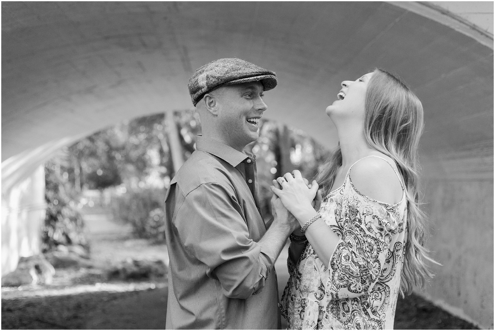 Engagement session couple laughing and holding hands while standing under a bridge.