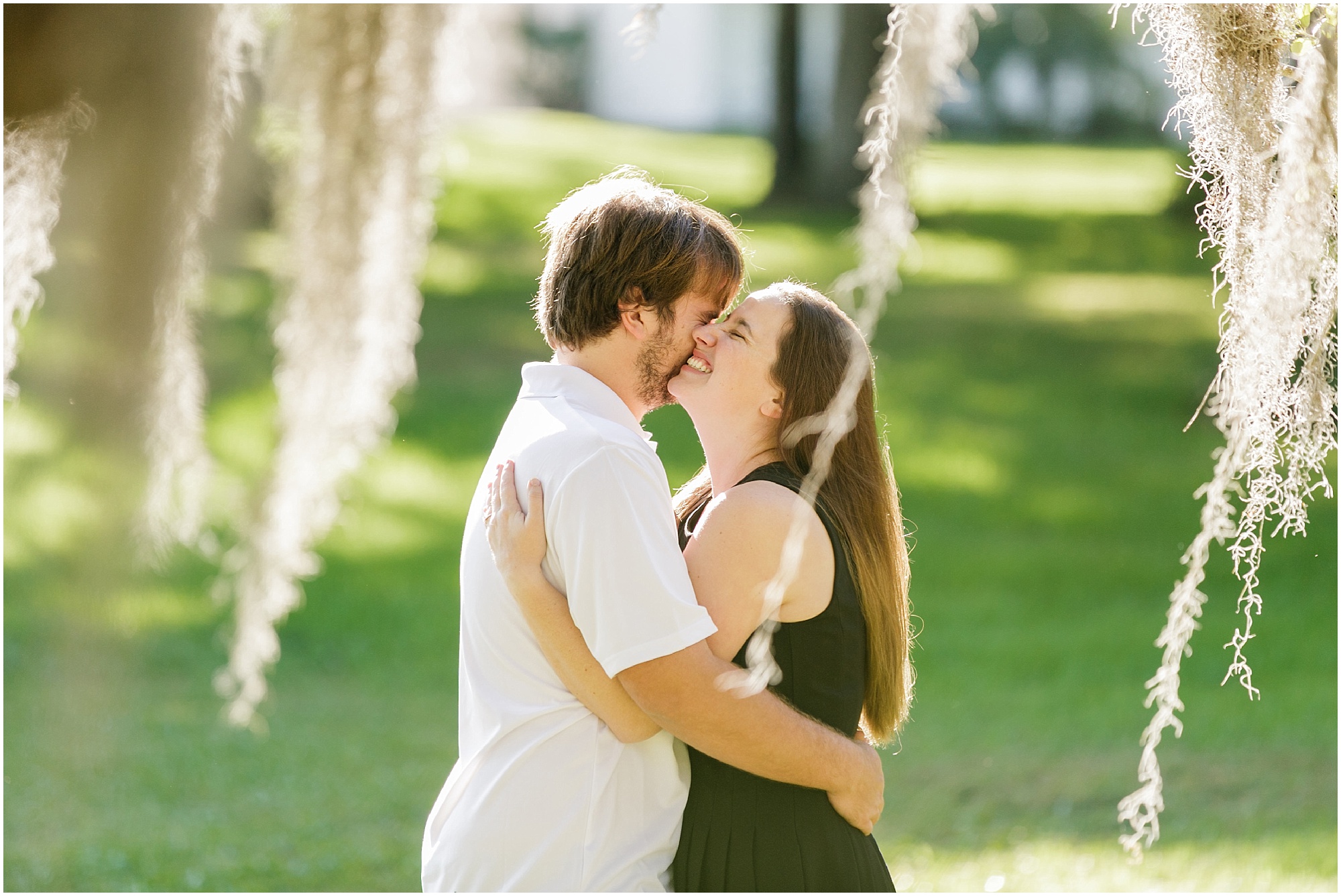 Couple snuggling and laughing under a moss tree in the sunlight. 