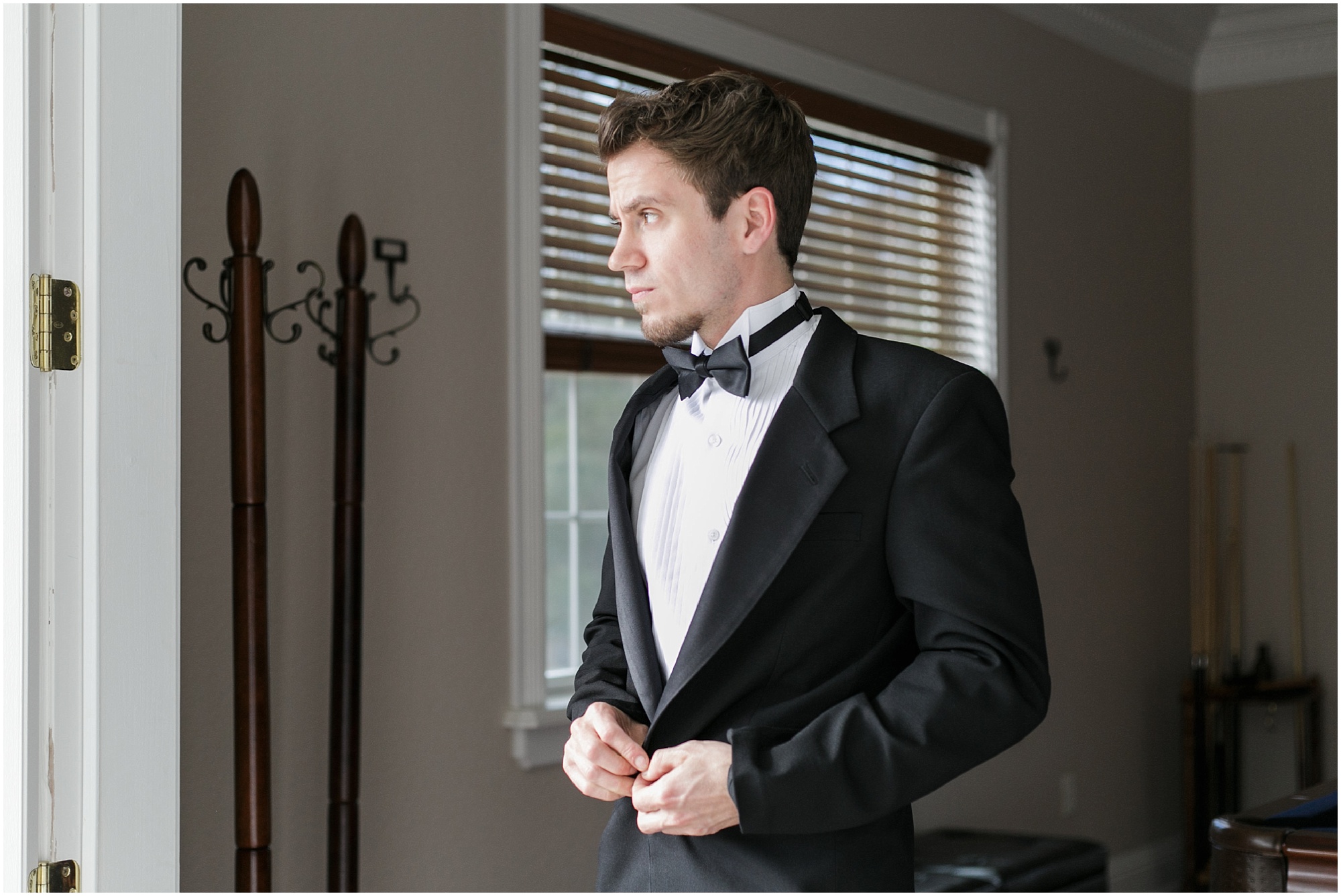 Groom looking out the window as he buttons up his tux jacket.