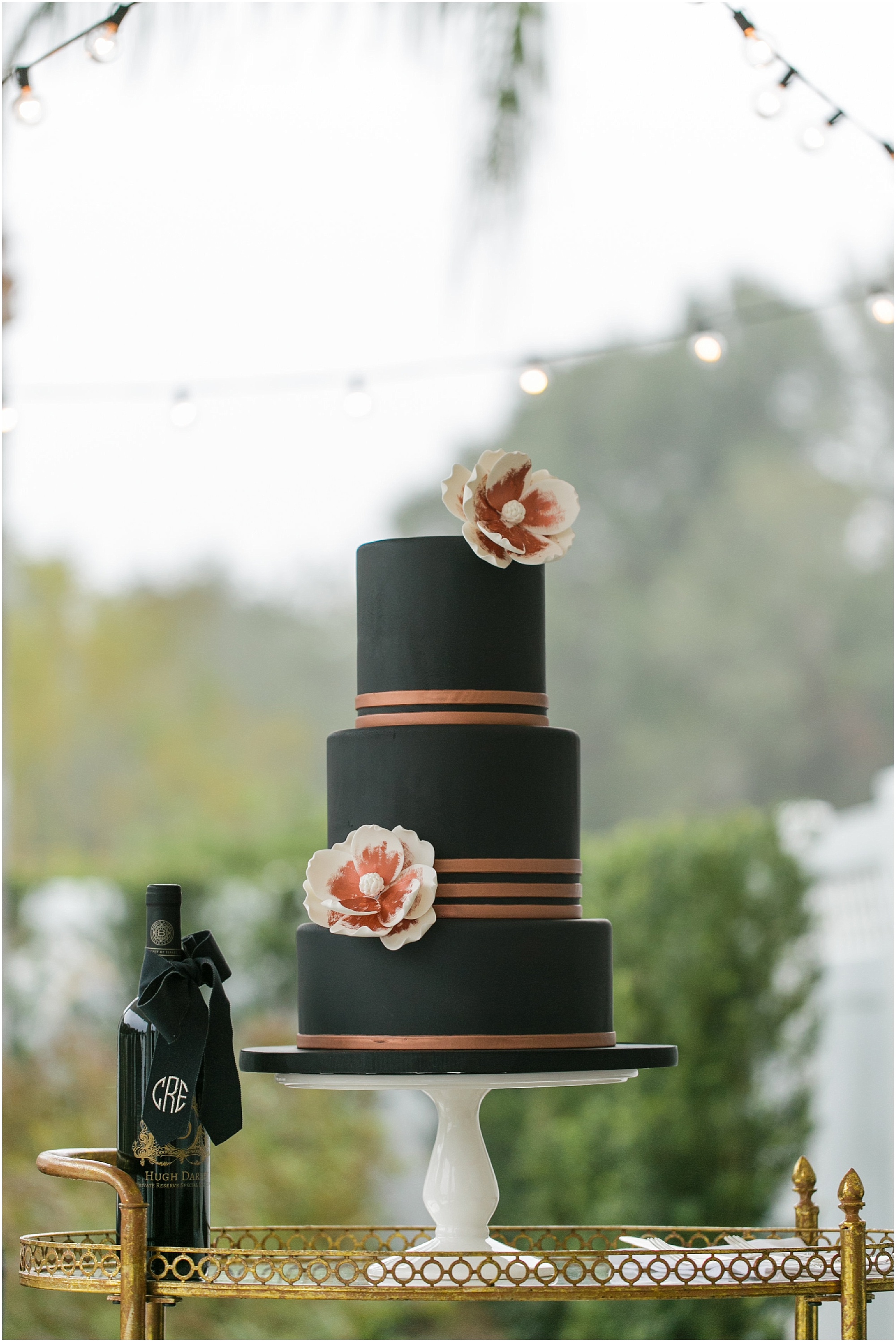 Wedding cake and wine bottle on the terrace with stringed lights in the background.