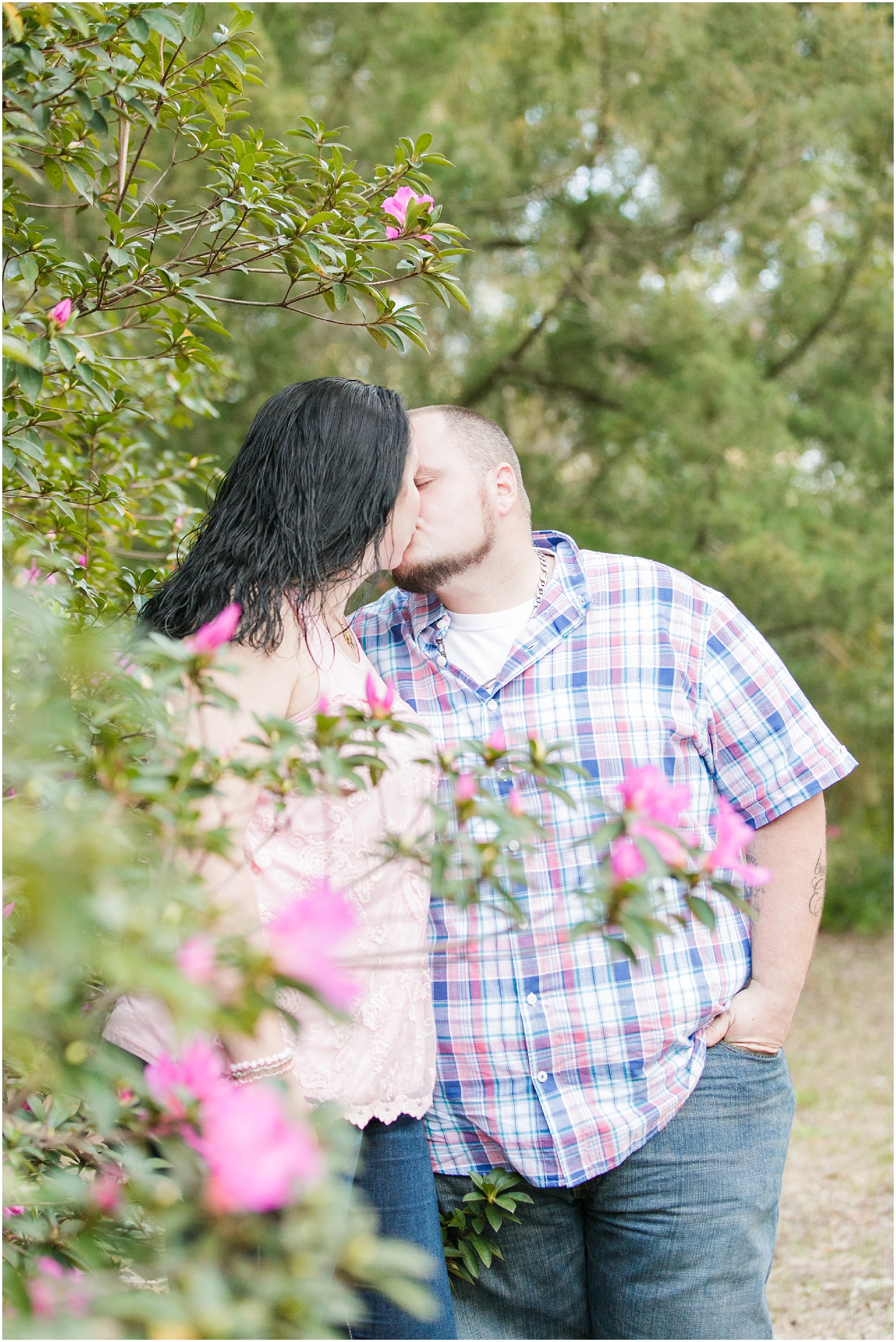 Couple share a kiss while standing near some bushes with light pink flowers.