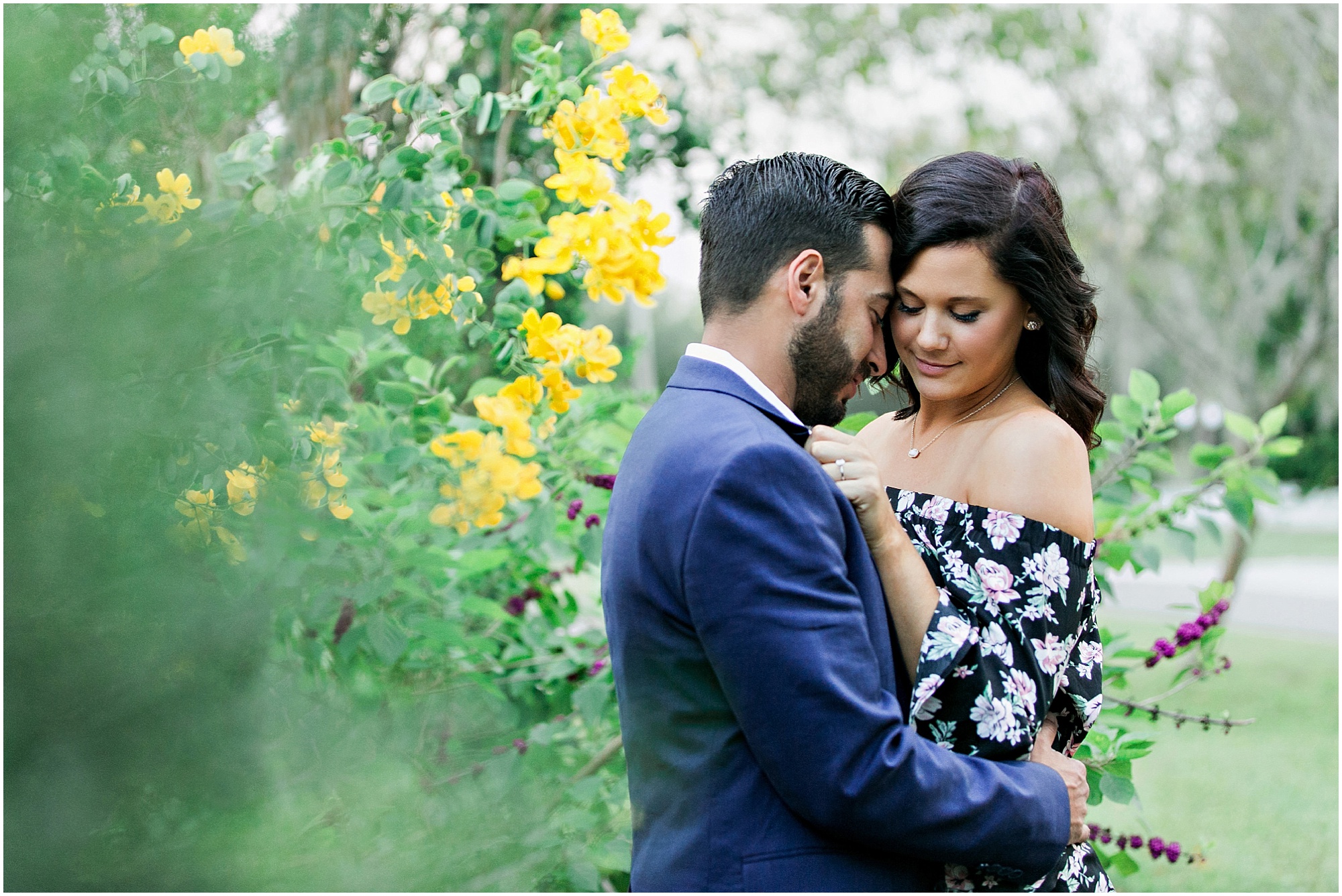 Engaged couple holding each other next to a wall of yellow flowers. 