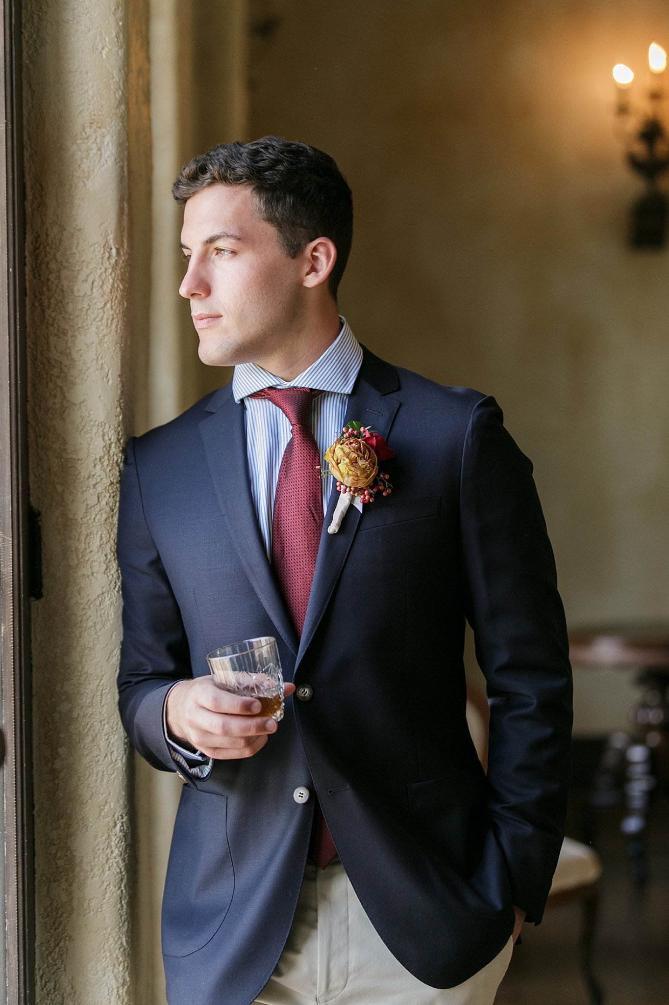 Groom looking out the window as he holds a drink in his hand. 