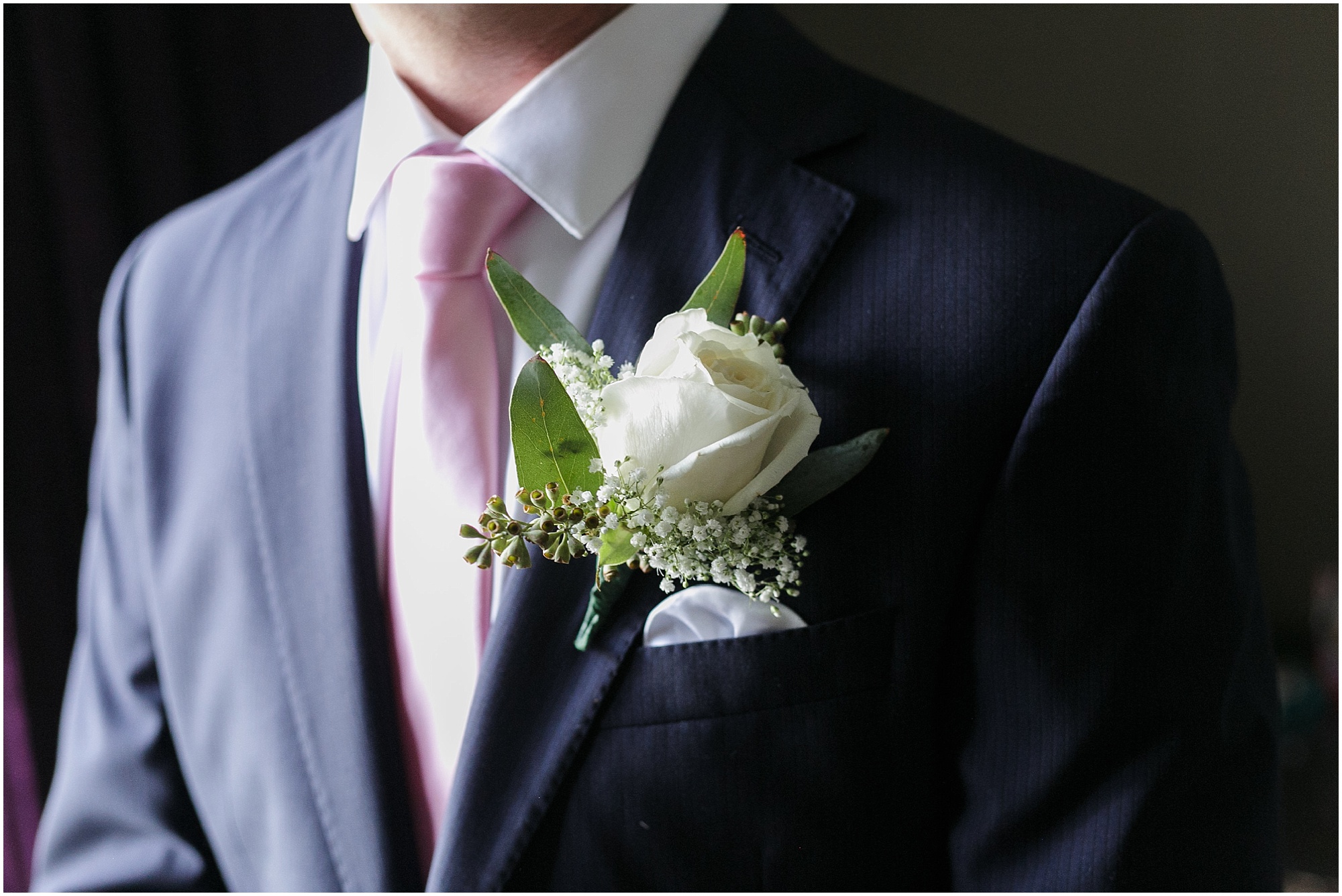 Close shot of the groom wearing a pink tie and a boutonniere made of a single while rose and baby's breath. 