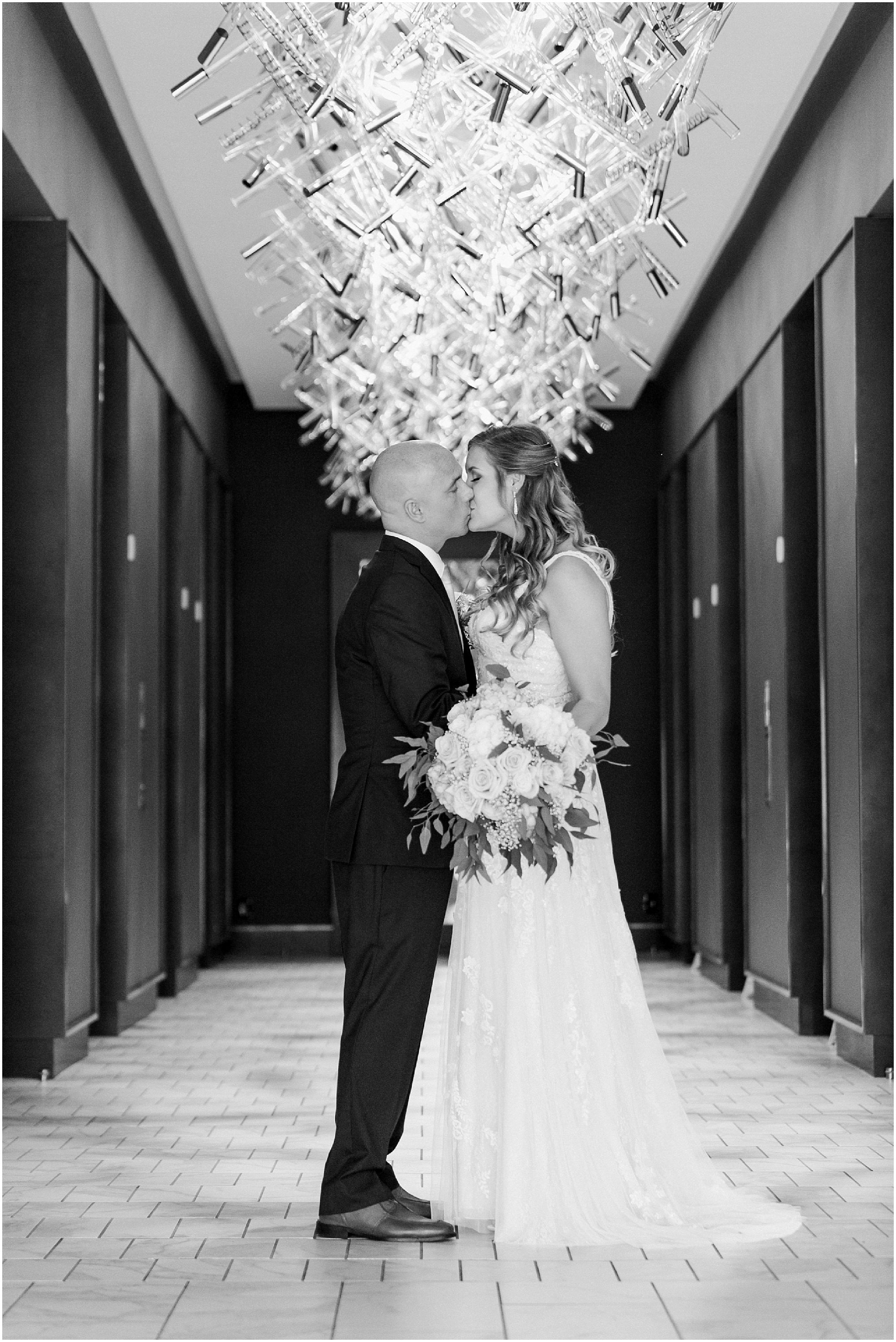 Black and white photo of the bride and groom kissing under a modern chandelier. 