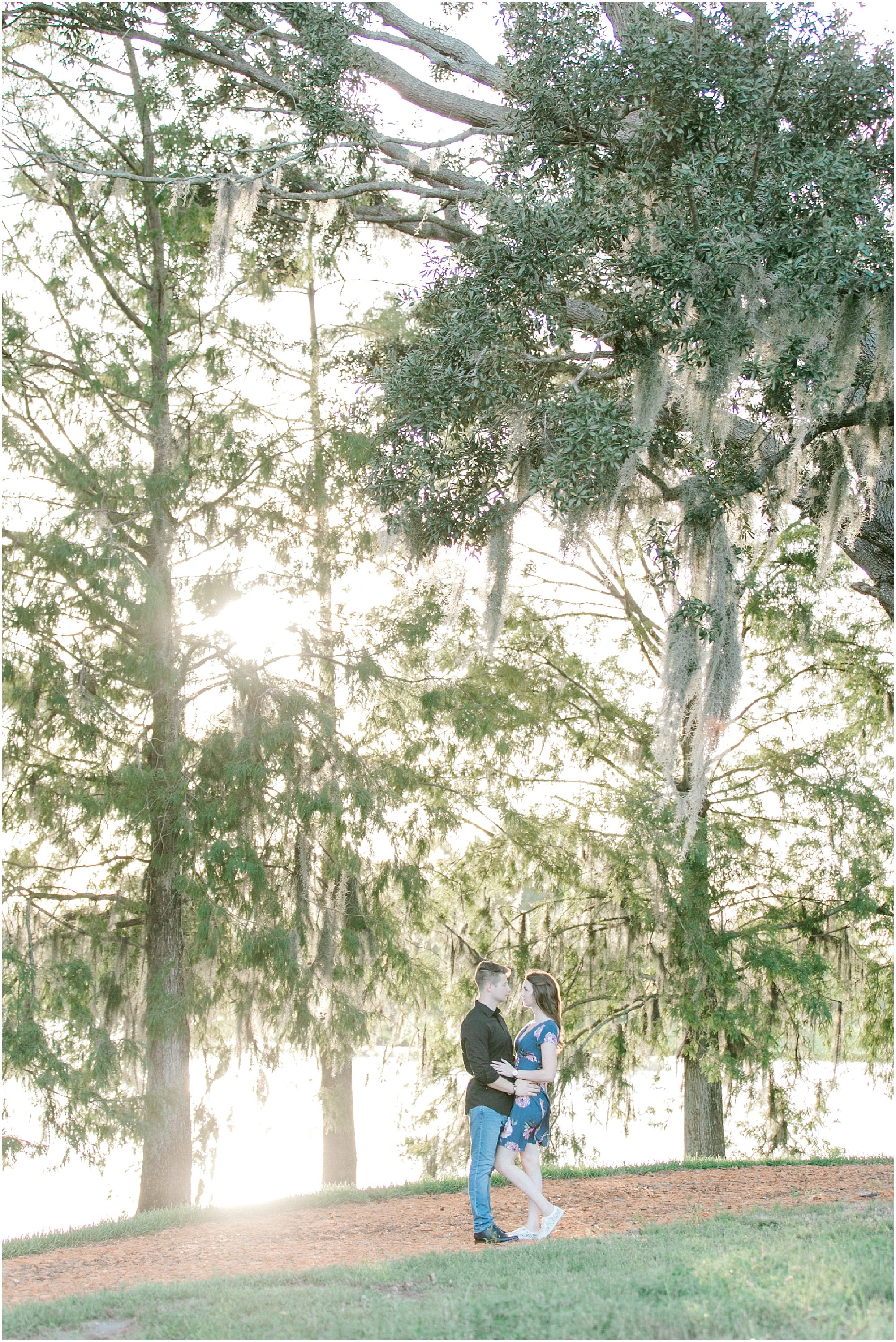 Couple looking at each other while standing under tall trees with the sun in the background