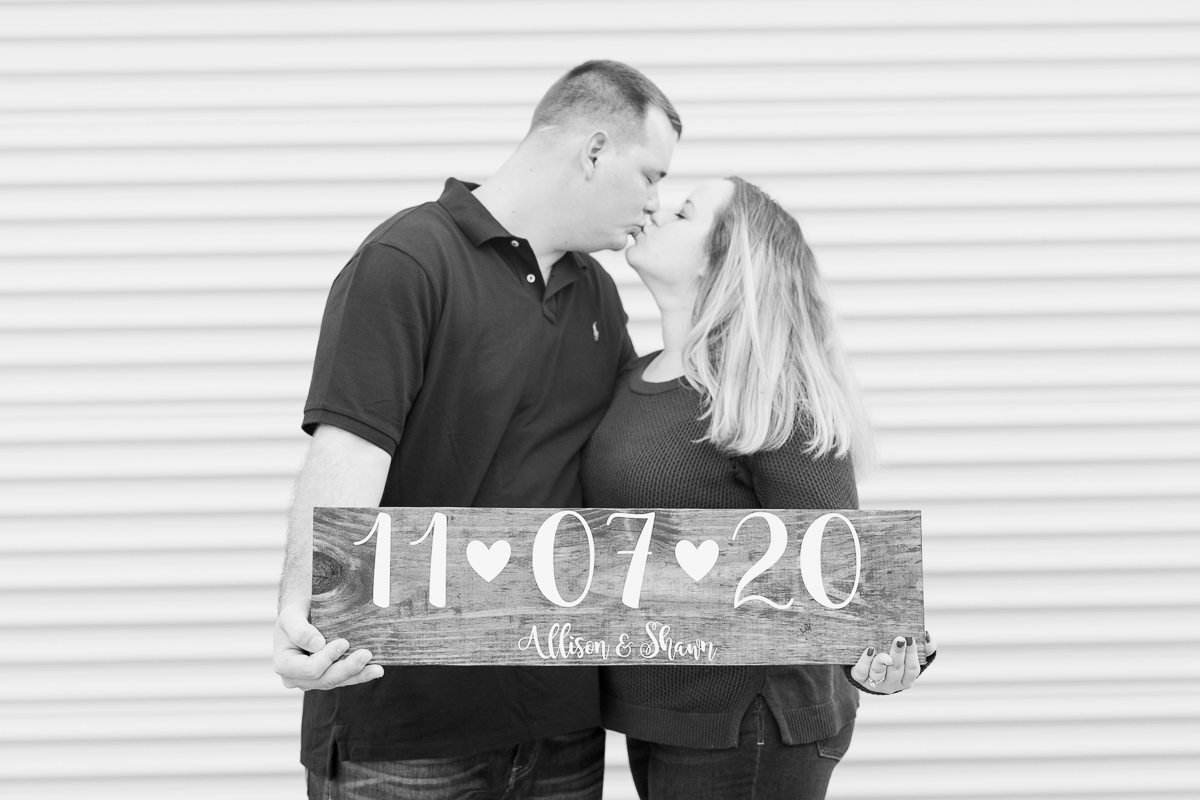 Black and white photo of couple kissing while holding wedding date sign