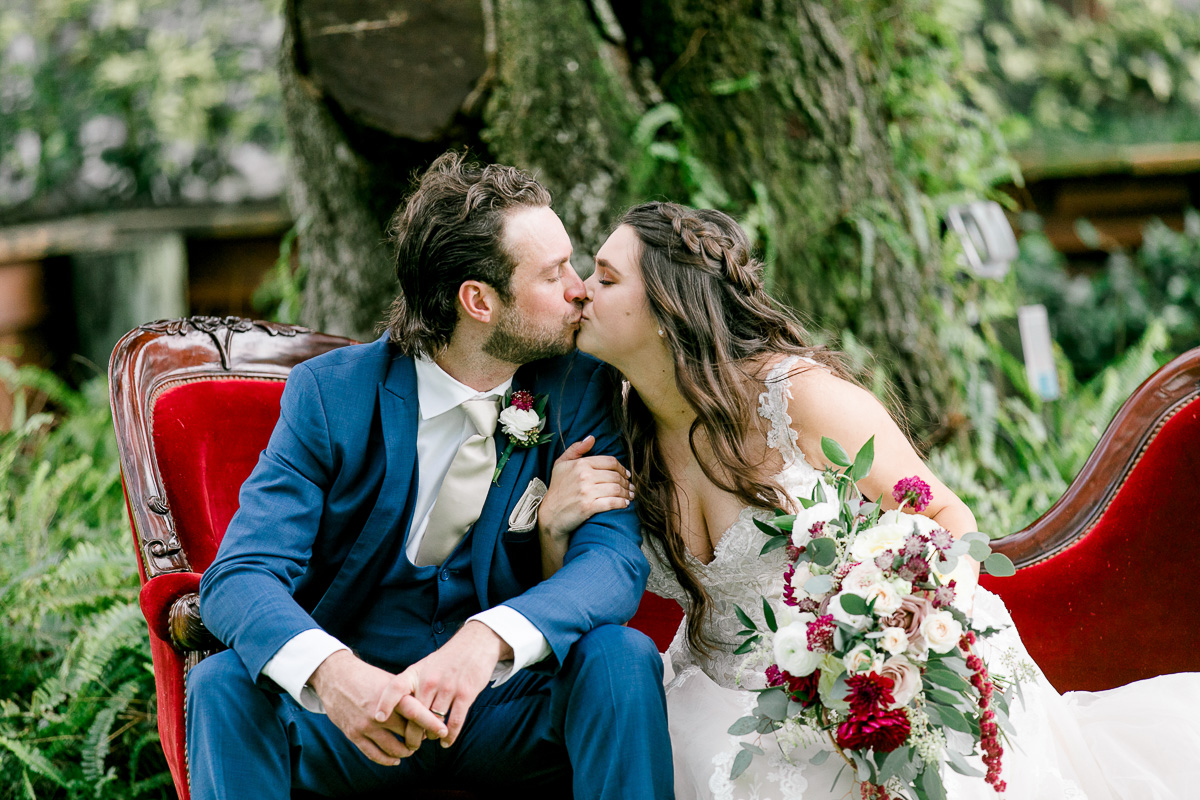 Bride and groom share a kiss while sitting on red vintage couch