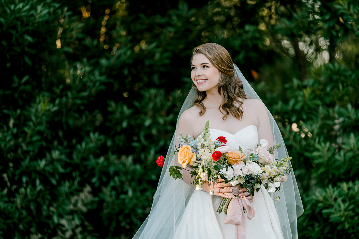 Bride with colorful bouquet standing in front of tall green bushes