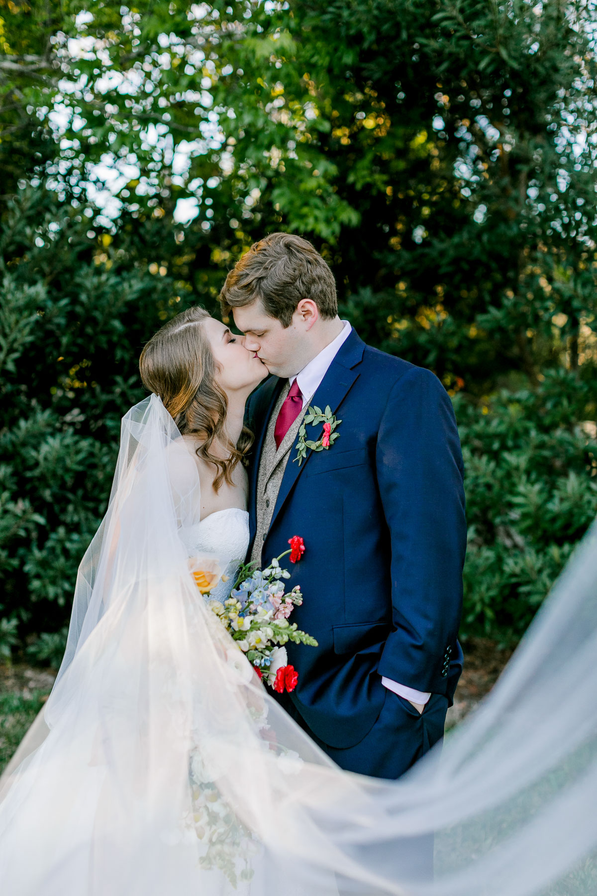 Bride and groom share a kiss in front of tall green bushes