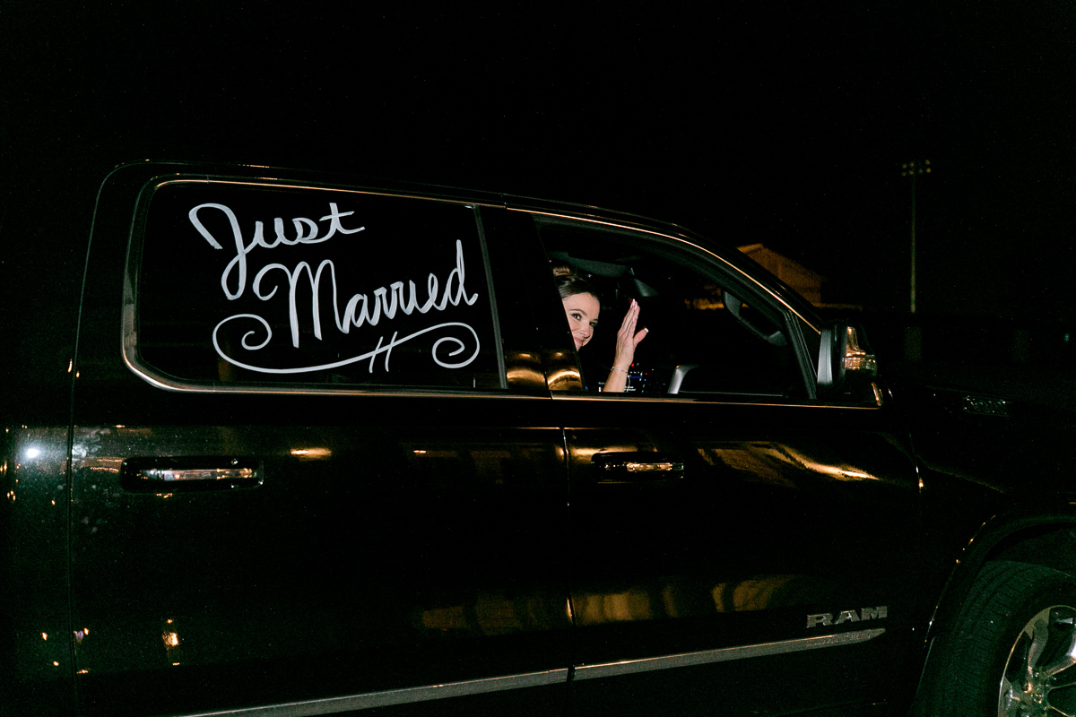 Newlyweds driving away from wedding in a truck that says "just married" on the window. 